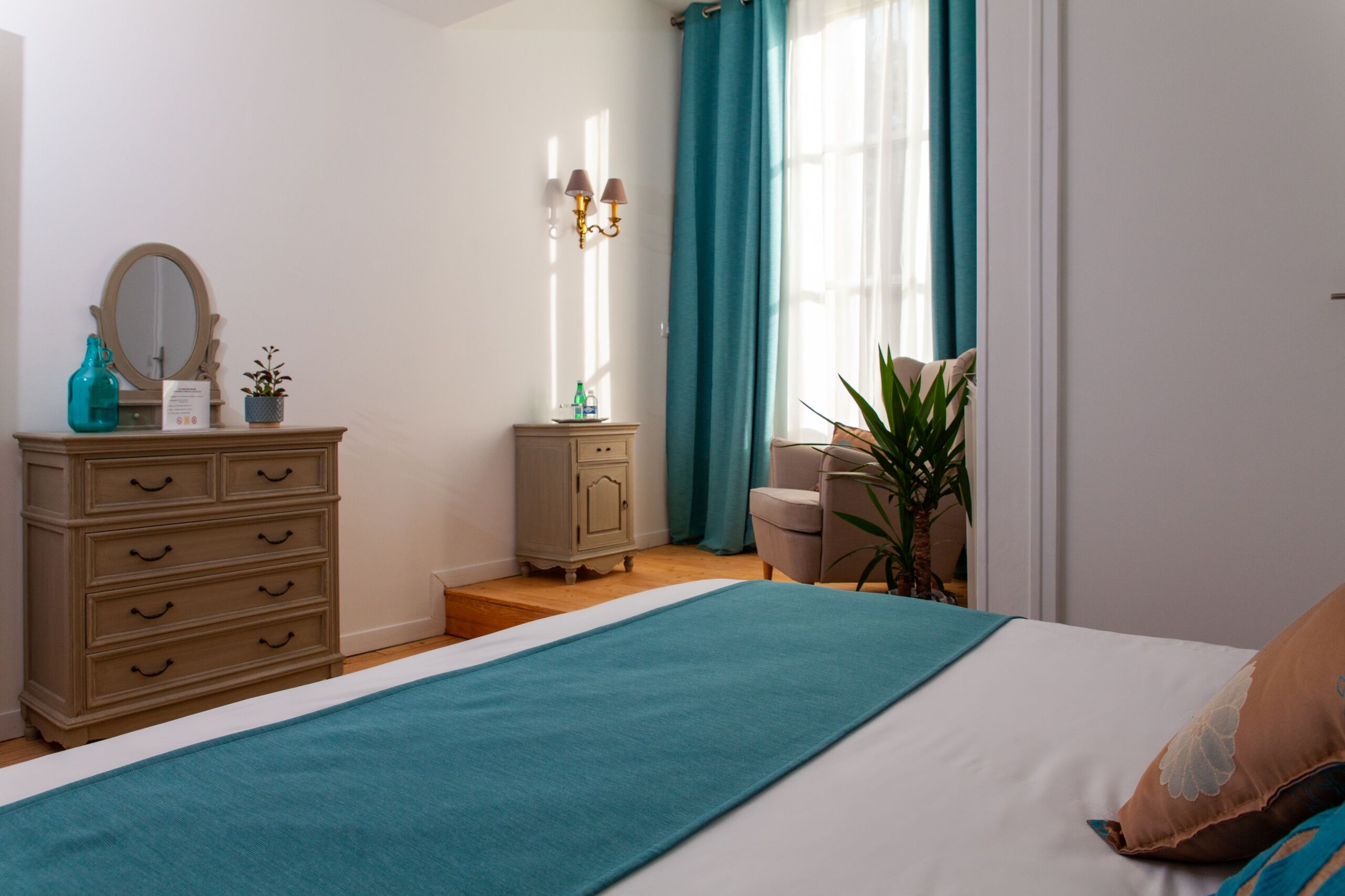Un dormitorio con cama, cómoda, mesita de noche, espejo, lámpara y ventana con cortinas.