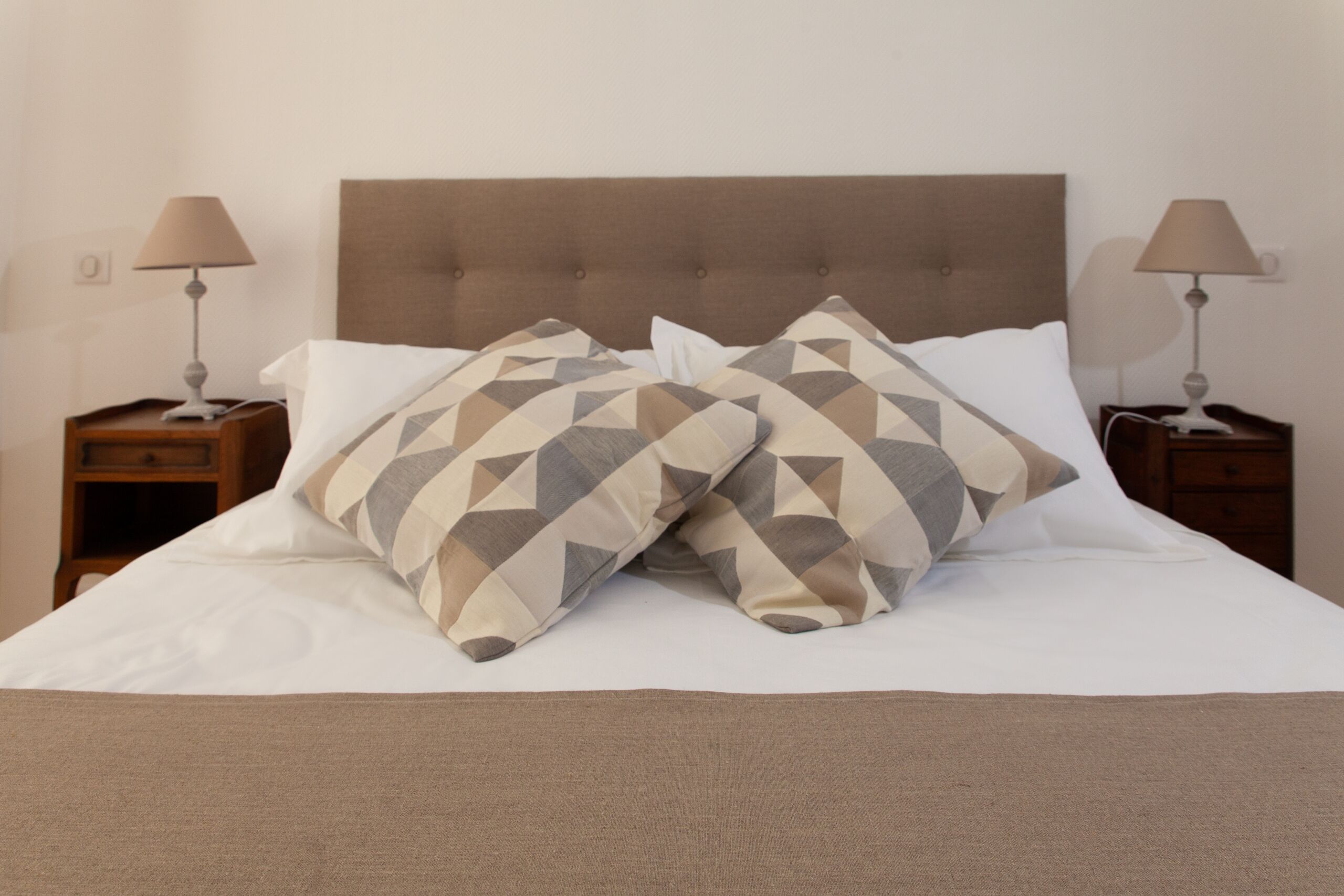 Una cama bien tendida con almohadillas estampadas, un cabecero acolchado y dos mesitas de noche con lámparas.