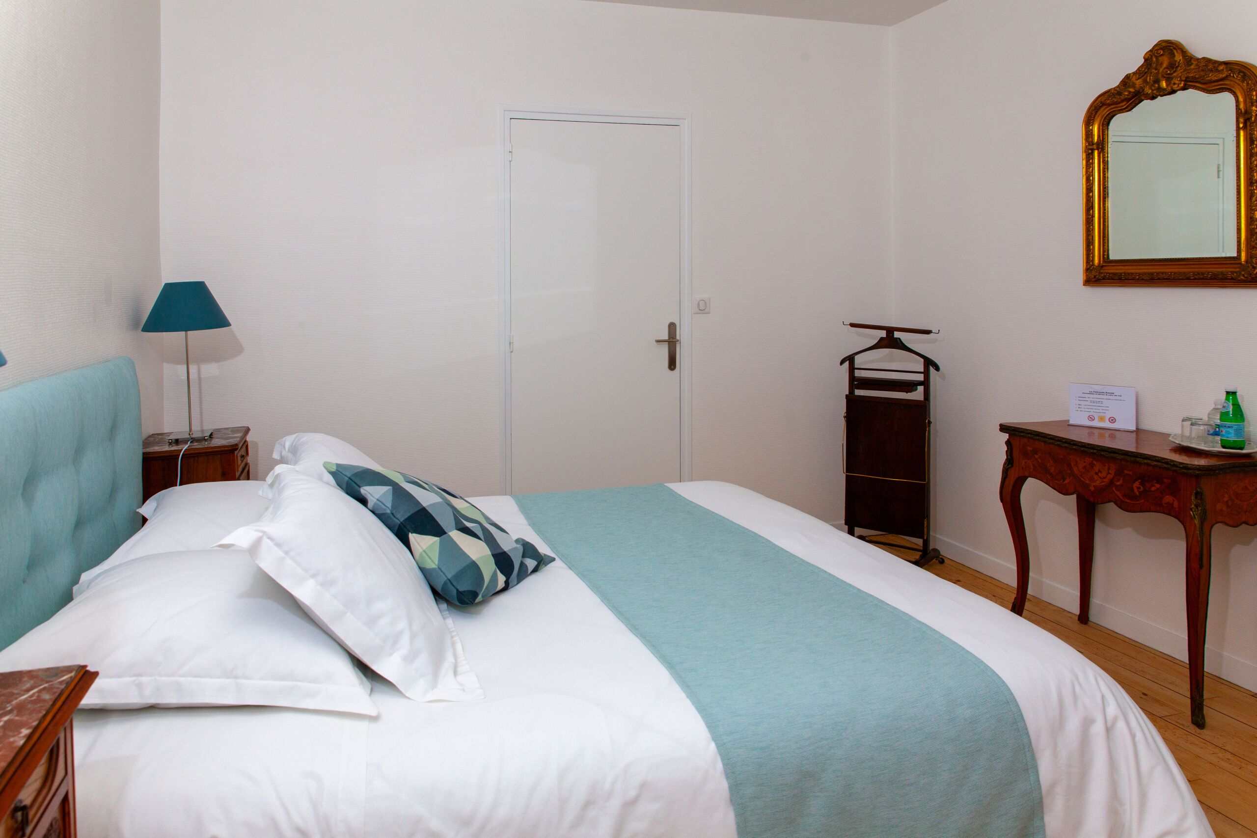 Ein ordentlich bezogenes Bett mit einer blauen und weißen Tagesdecke, ein Nachttisch mit einer Lampe, eine Holzkommode und ein Spiegel an der Wand.