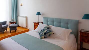 Een netjes opgemaakt bed met een blauw hoofdeinde, een nachtkastje met een lamp, een stoel en een klein tafeltje met een plant.