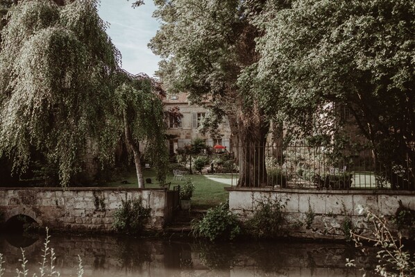 Un pont de pedra sobre una massa d'aigua tranquil·la, envoltat de vegetació exuberant i un edifici de pedra amb una tenda vermella al fons.