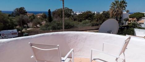 A rooftop terrace with two white chairs, a white wall, and a view of the sea and buildings in the background.
