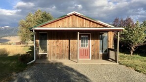 A wooden cabin with a red door and a small porch, surrounded by a gravel driveway and grassy areas. - Cozy cottage nestled in Paradise Valley surrounded by mountain views! (Livingston)