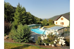 Piscina con cerramiento de vidrio, rodeada de abundante vegetación y una casa al fondo.