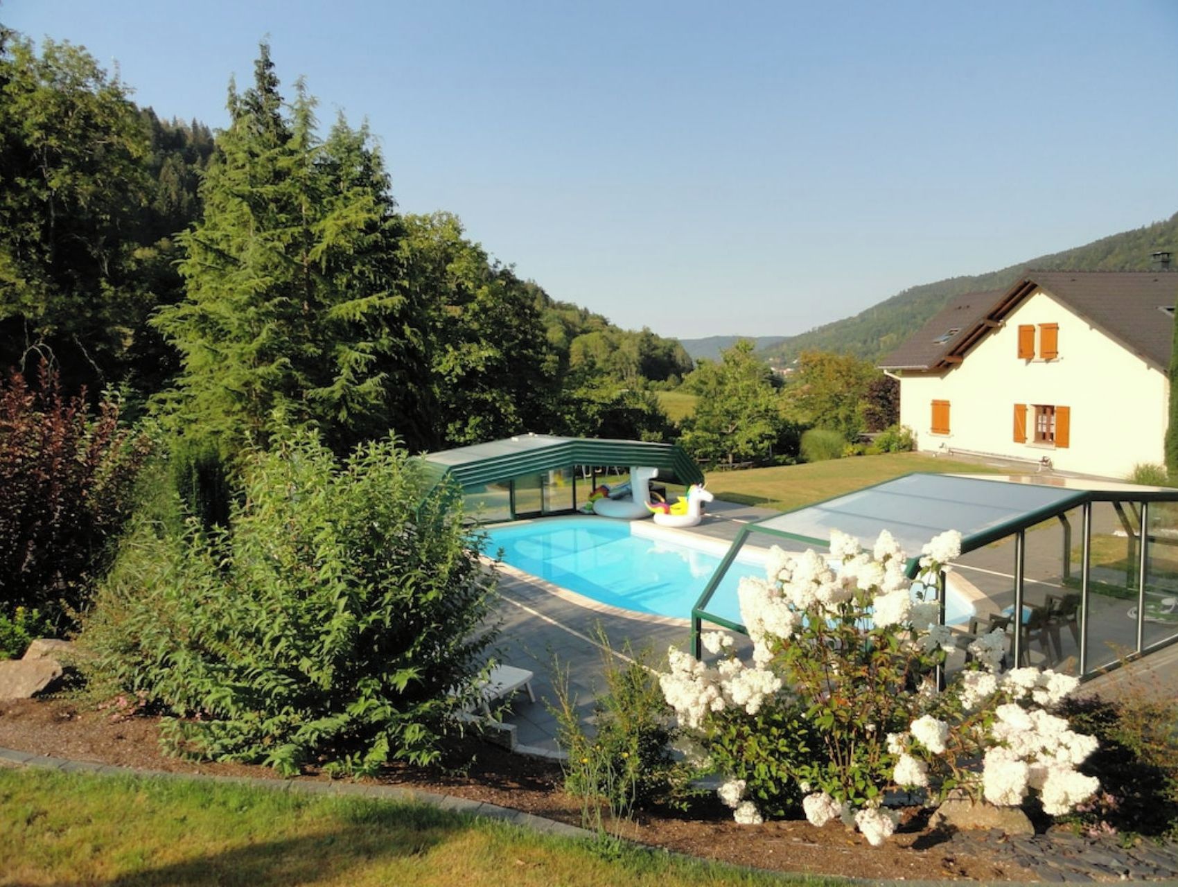 A swimming pool with a glass enclosure, surrounded by lush greenery and a house in the background.