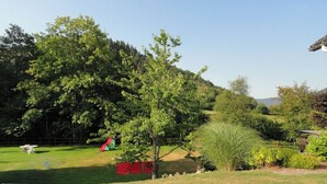 Un exuberante jardín con diversas especies de árboles y arbustos, una pequeña estructura roja para juegos y una zona con césped.