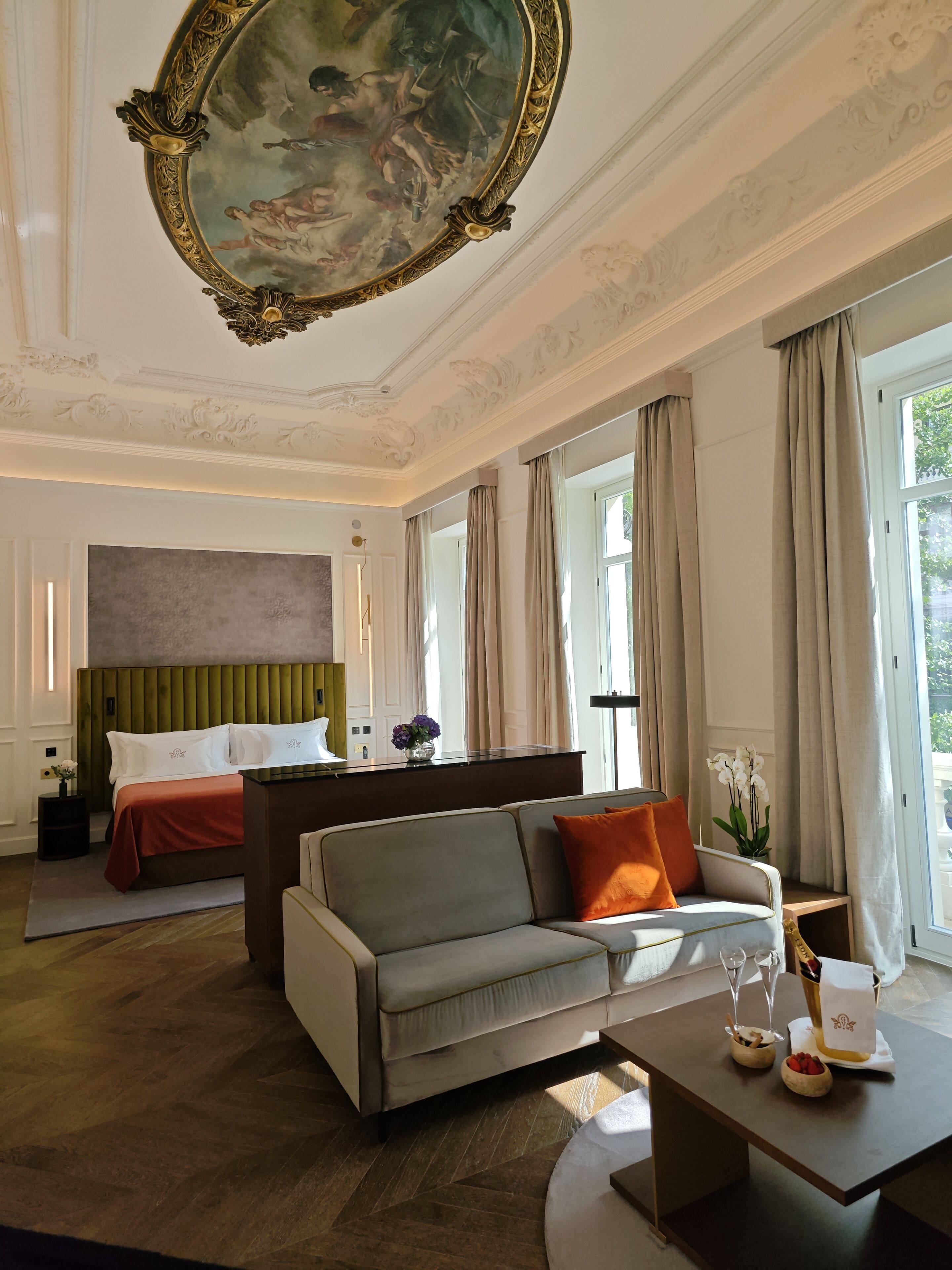 A hotel room with a bed, a sofa, a dining table with glasses and a vase, and a large painting on the ceiling.