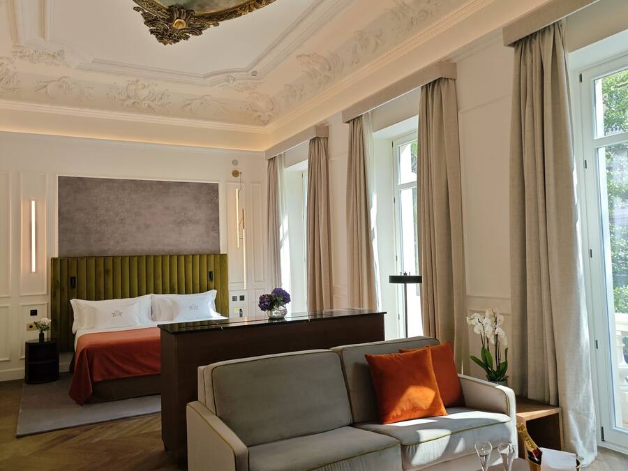 A hotel room with a bed, a sofa, a dining table with glasses and a vase, and a large painting on the ceiling.