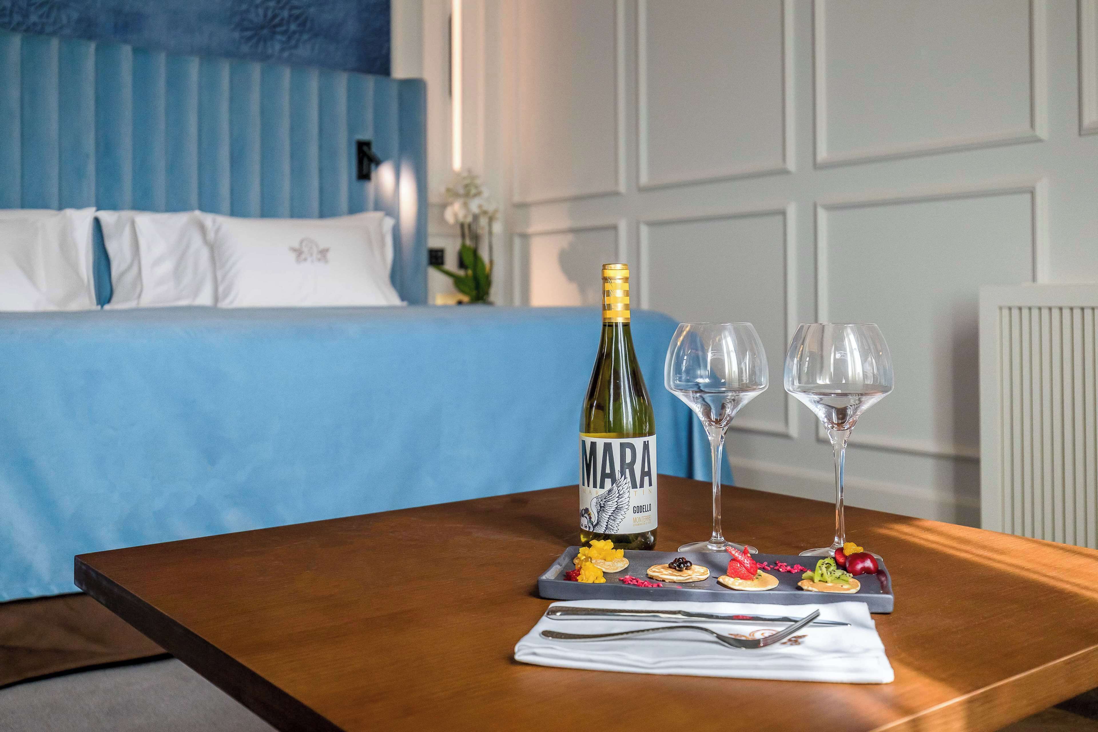 Habitación de hotel con cama, una mesa de madera con una botella de vino, dos copas y una bandeja con comida.