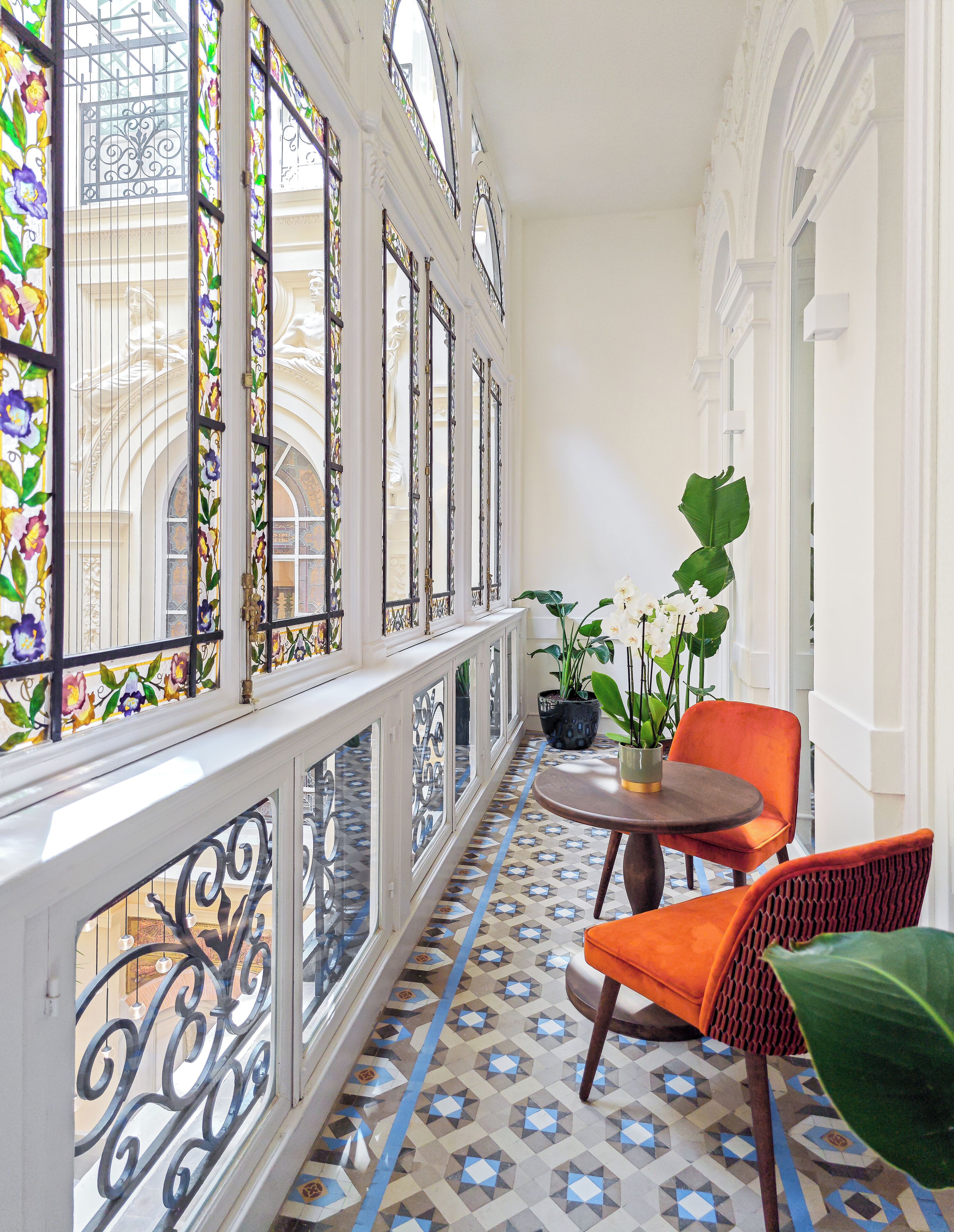 Un pasillo con vidrieras decorativas, suelo de baldosas estampadas y una zona de estar con sillas y una mesa.