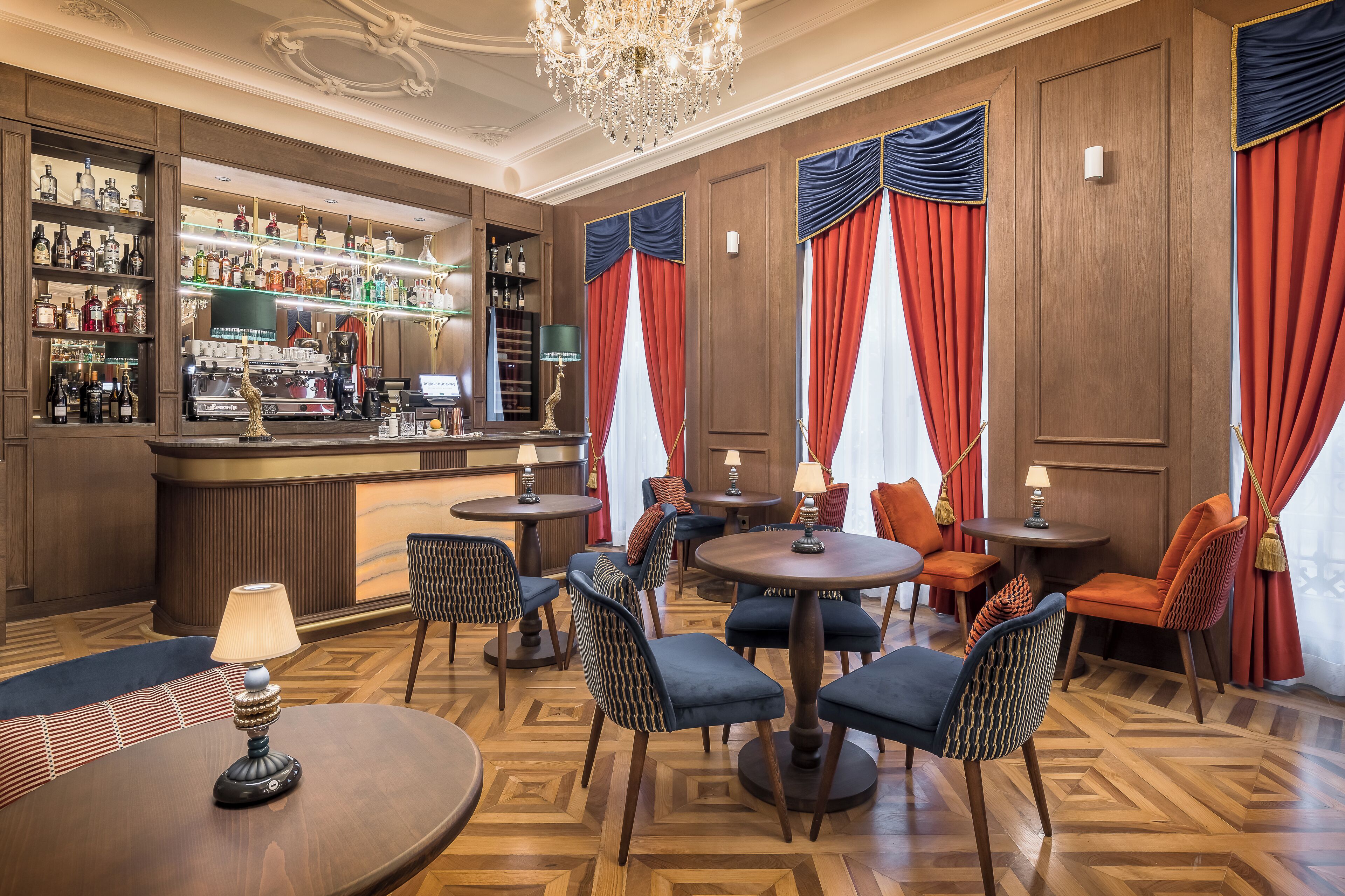 A bar area with wooden paneling, a chandelier, and a variety of liquor bottles. There are multiple tables and chairs arranged for seating.