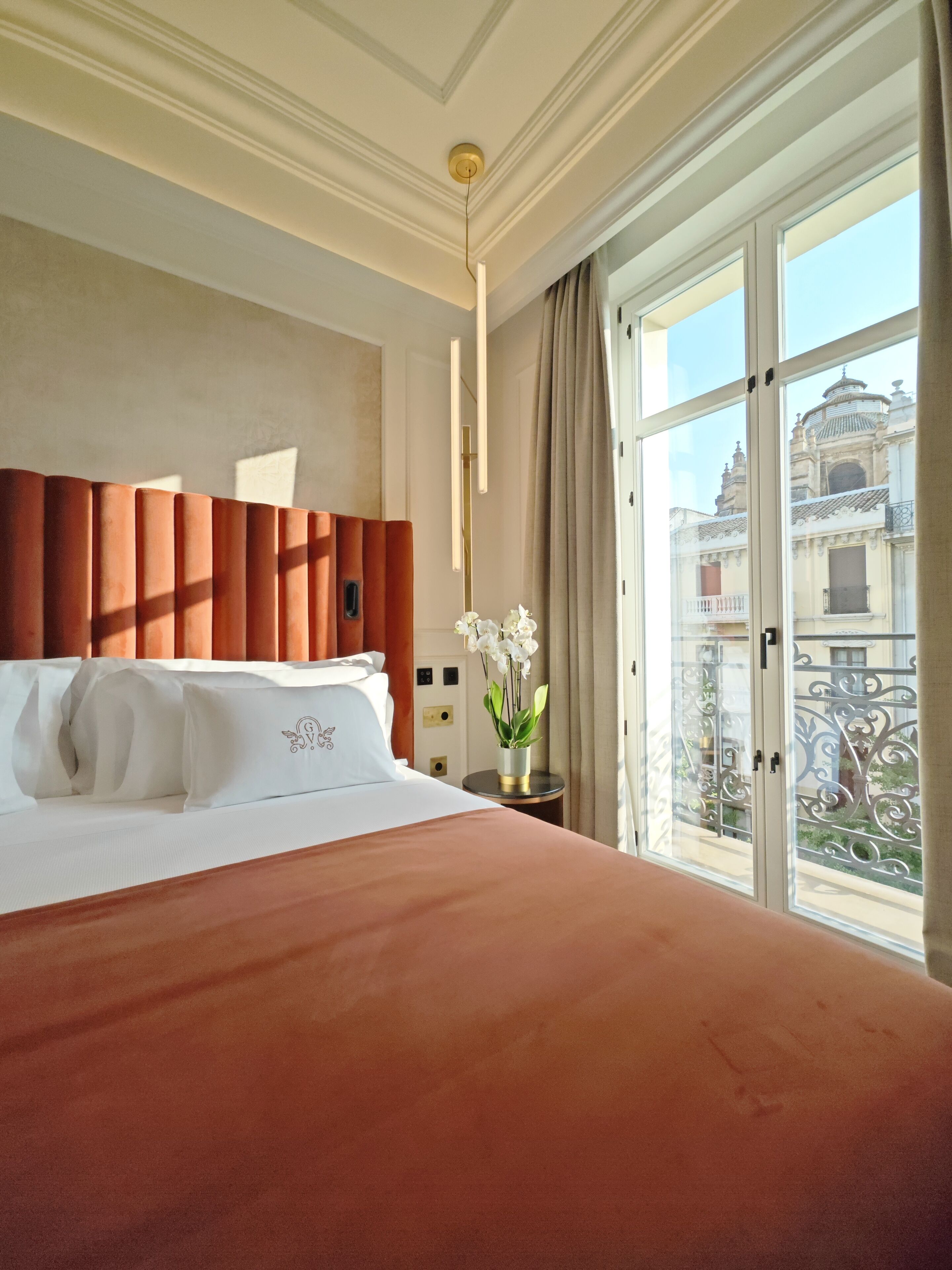 Une chambre d’hôtel avec un grand lit, une vue sur un immeuble par la fenêtre et une table de chevet ornée d’un vase de fleurs.
