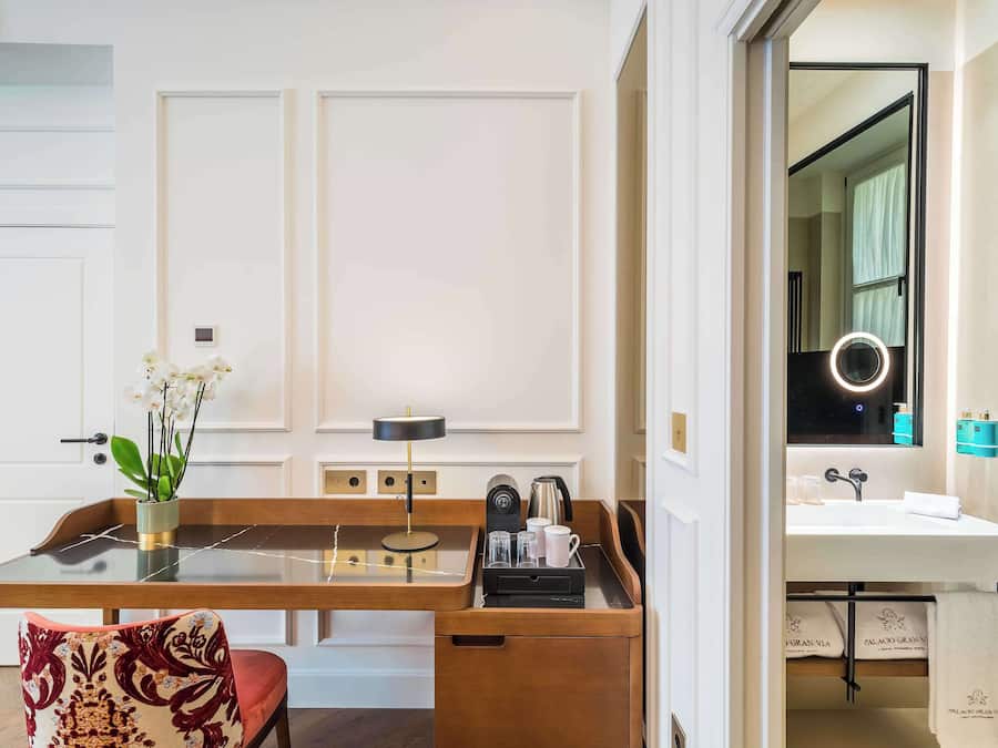 Une chambre d’hôtel avec un bureau en bois, une chaise dotée d’un coussin à motifs, un vase avec des fleurs, une lampe, une machine à café et un plateau avec une tasse et des verres.
