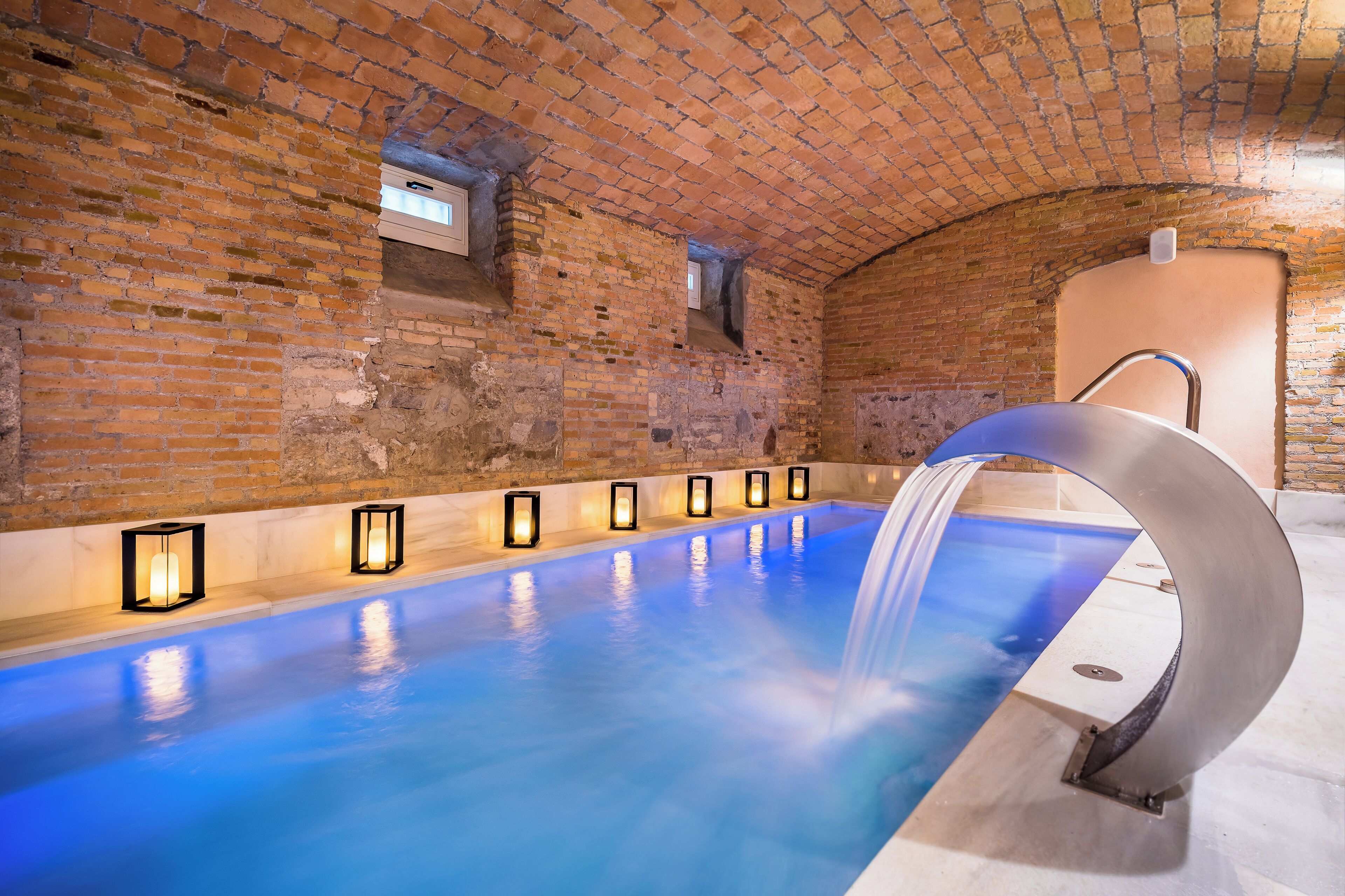 A swimming pool with a water feature, surrounded by brick walls and illuminated by recessed lighting.