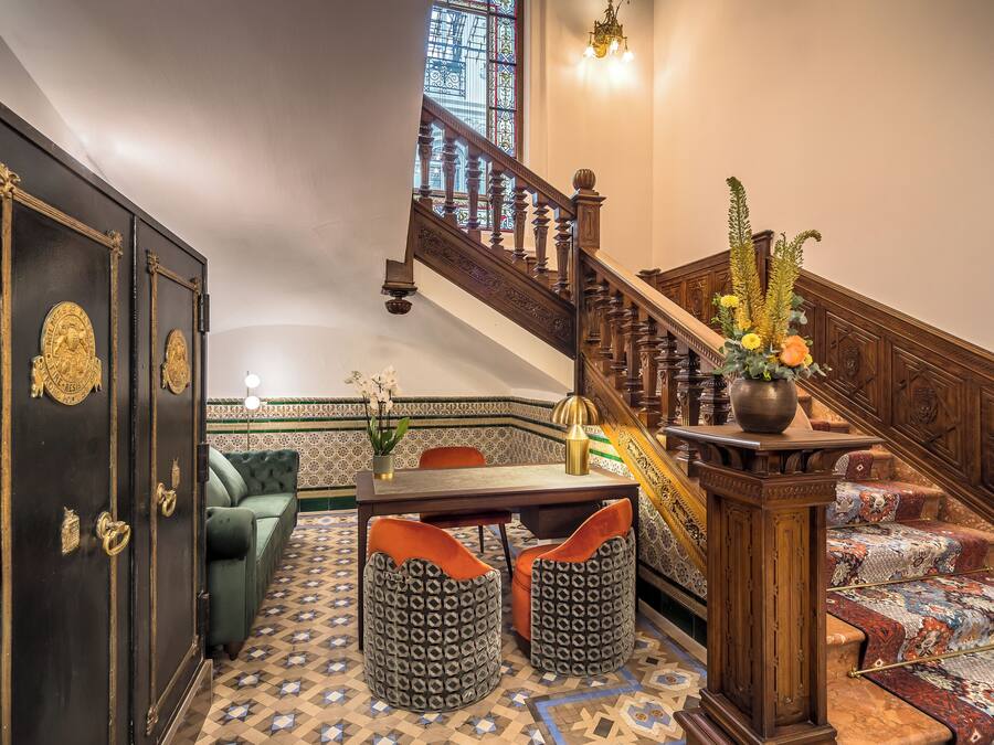 Una escalera con detalles de madera decorativos, un sofá verde, otomanas estampadas y una mesa de comedor con sillas.