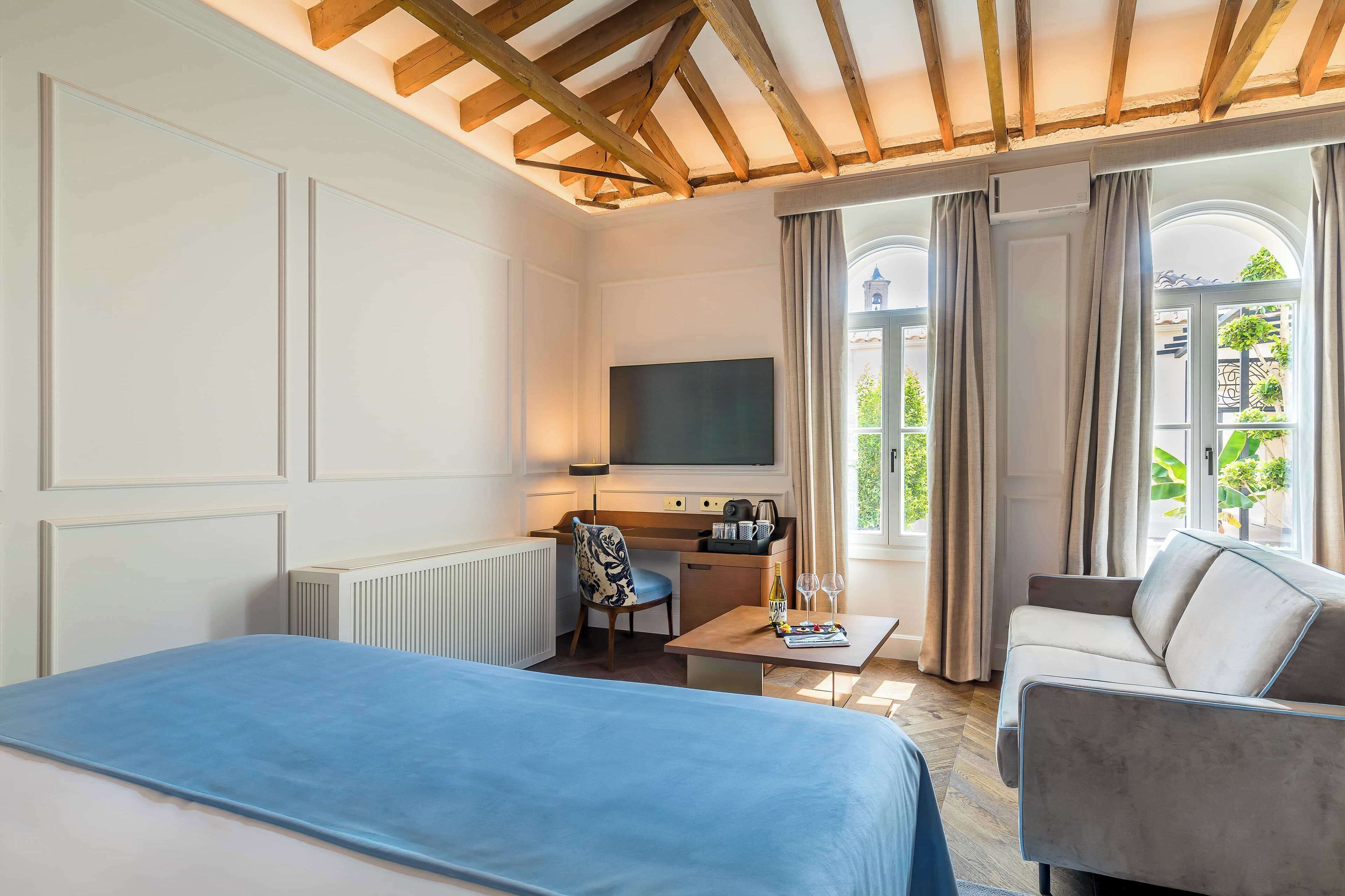 Habitación de hotel con una cama, un escritorio con silla, un sofá, un televisor y una ventana con cortinas.