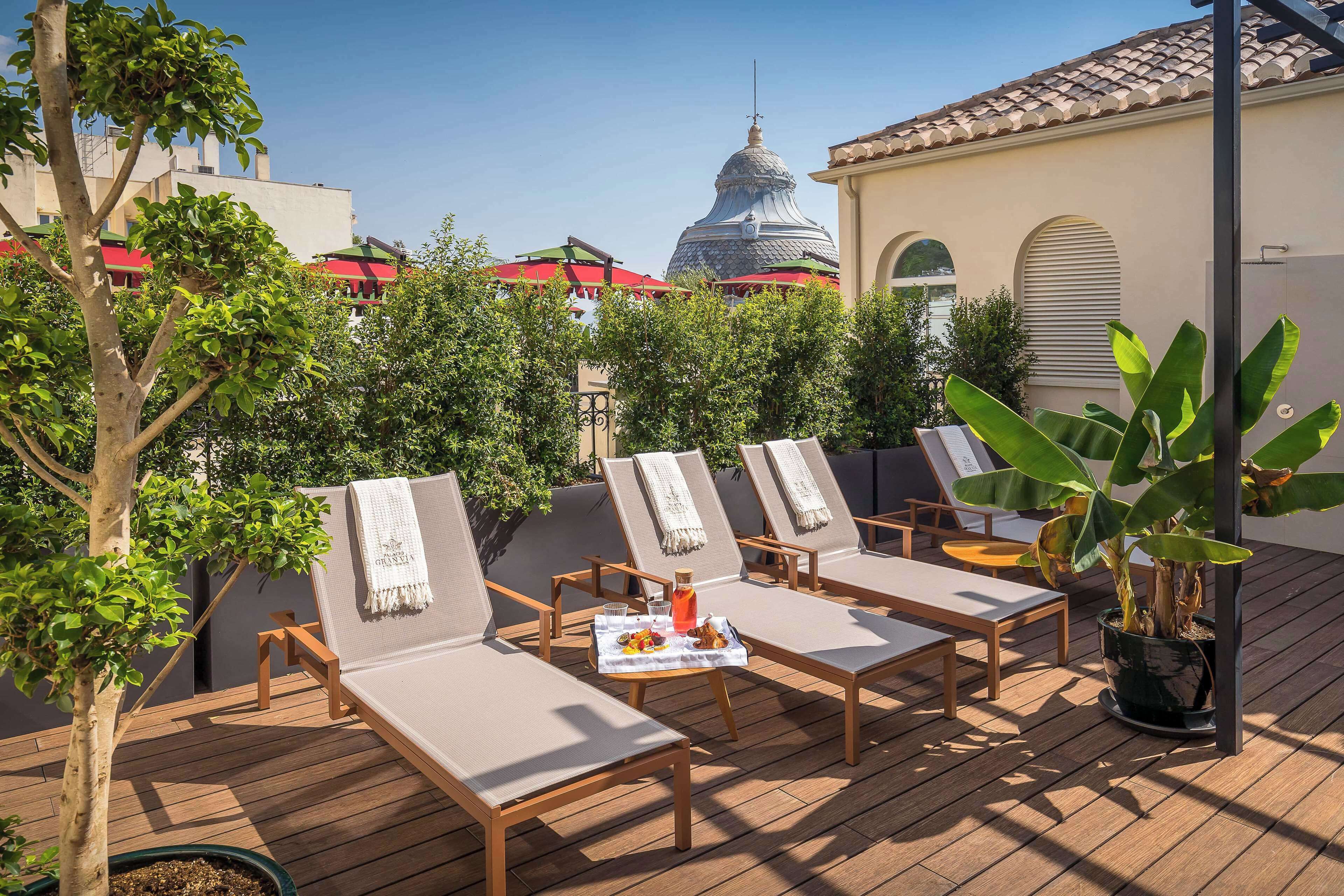 Terraza en la azotea con sillones de descanso, una mesa con bebidas y aperitivos, y vistas a un edificio con cúpula y tejados rojos.