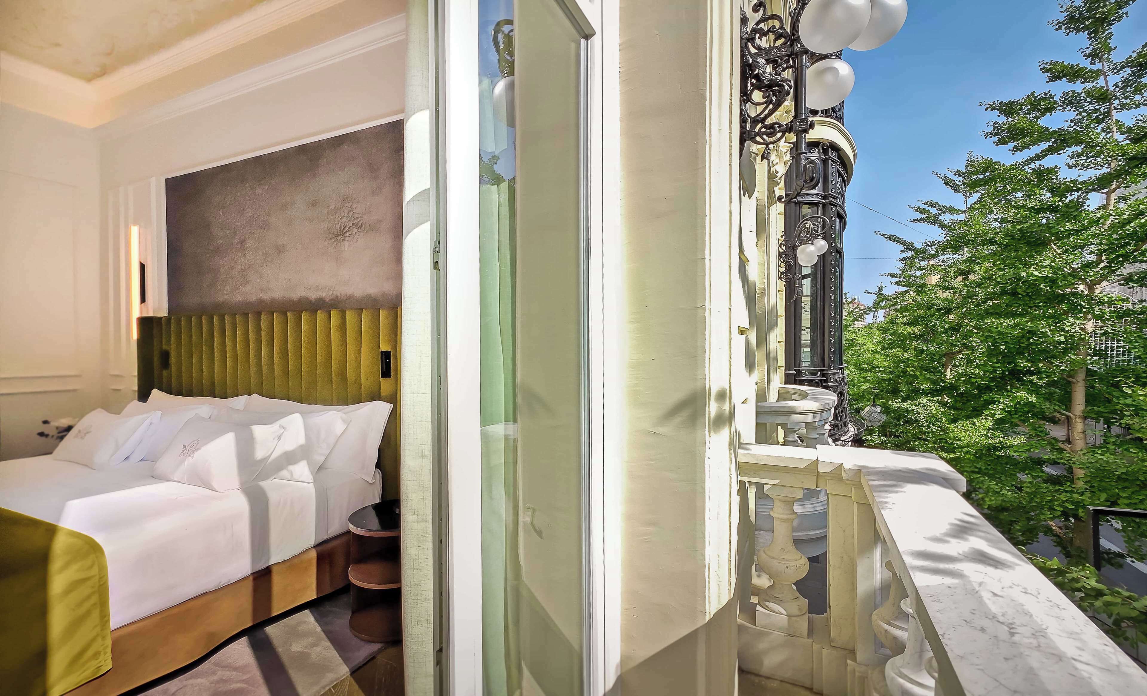 Habitación de hotel con cama, cabecera verde, balcón con barandilla de hierro forjado y vista a árboles.