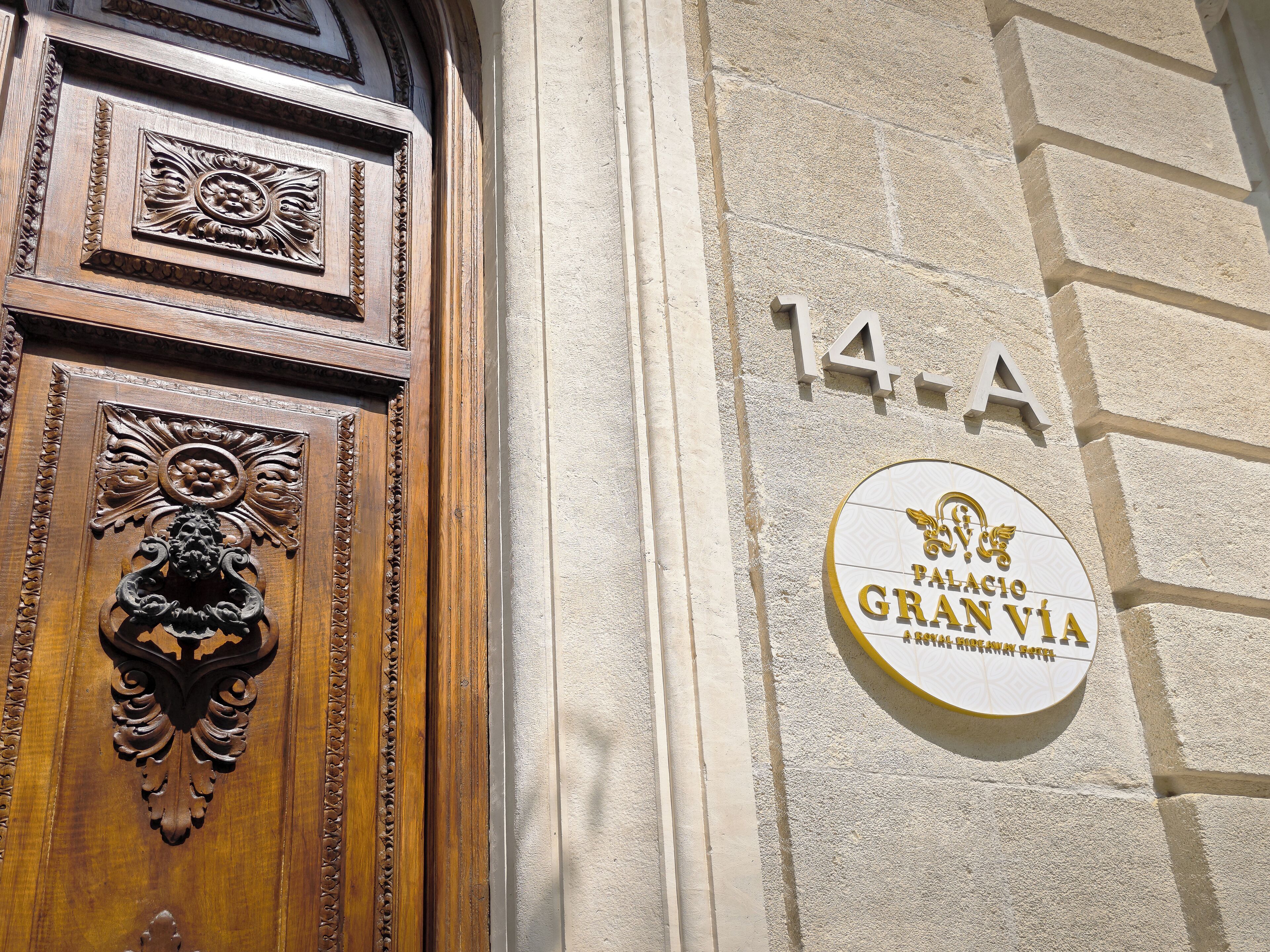 A building entrance with a wooden door featuring intricate carvings and a brass knocker. A sign above the door displays the number "14-A" and the name "Palacio Gran Vía".
