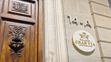 A building entrance with a wooden door featuring intricate carvings and a brass knocker. A sign above the door displays the number "14-A" and the name "Palacio Gran Vía".