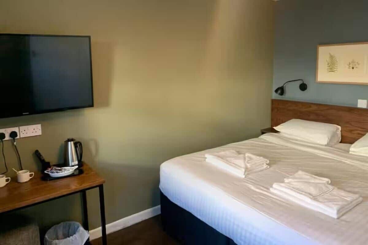 A neatly made bed with white linens, a bedside table with a coffee maker, and a flat-screen TV mounted on the wall.