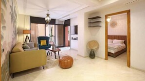 A modern living room with a sofa, a round ottoman, a coffee table, and a blue armchair. There is a bed with a wooden headboard in the background. - Jolie Appart-centre Gueliz (Marrakesh)