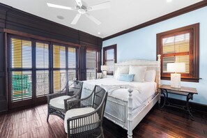Ein Schlafzimmer mit einem großen Bett, zwei Sesseln, einem Deckenventilator und mehreren Fenstern mit Jalousien.