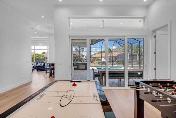 Une pièce spacieuse équipée d’un baby-foot, d’une table de jeu de palet et d’une table de billard. Une grande porte-fenêtre coulissante donne accès à un patio extérieur avec une piscine et des espaces pour s’asseoir.