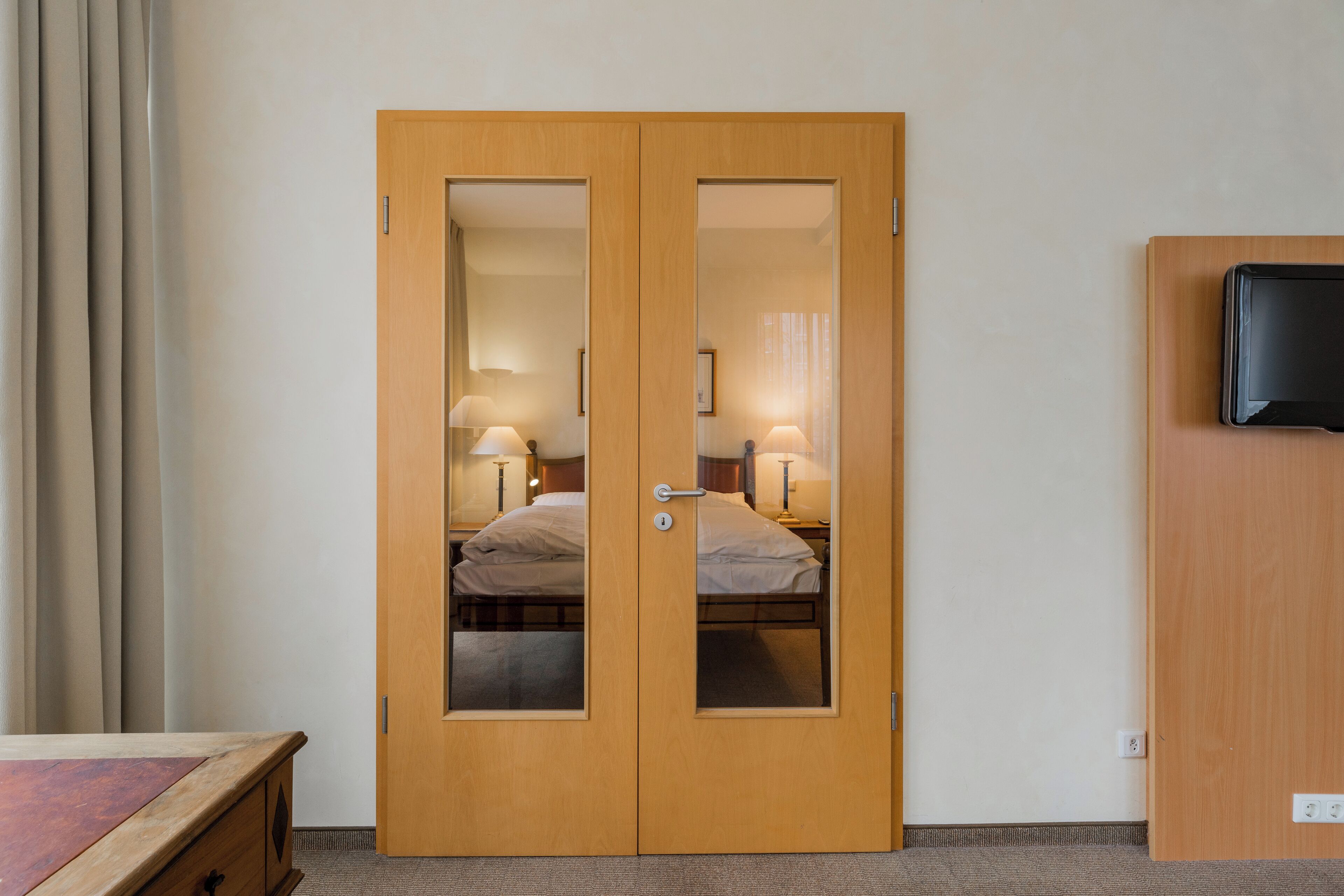 A hotel room with a wooden double door, a bed with white linens, a bedside table, and a television mounted on the wall.