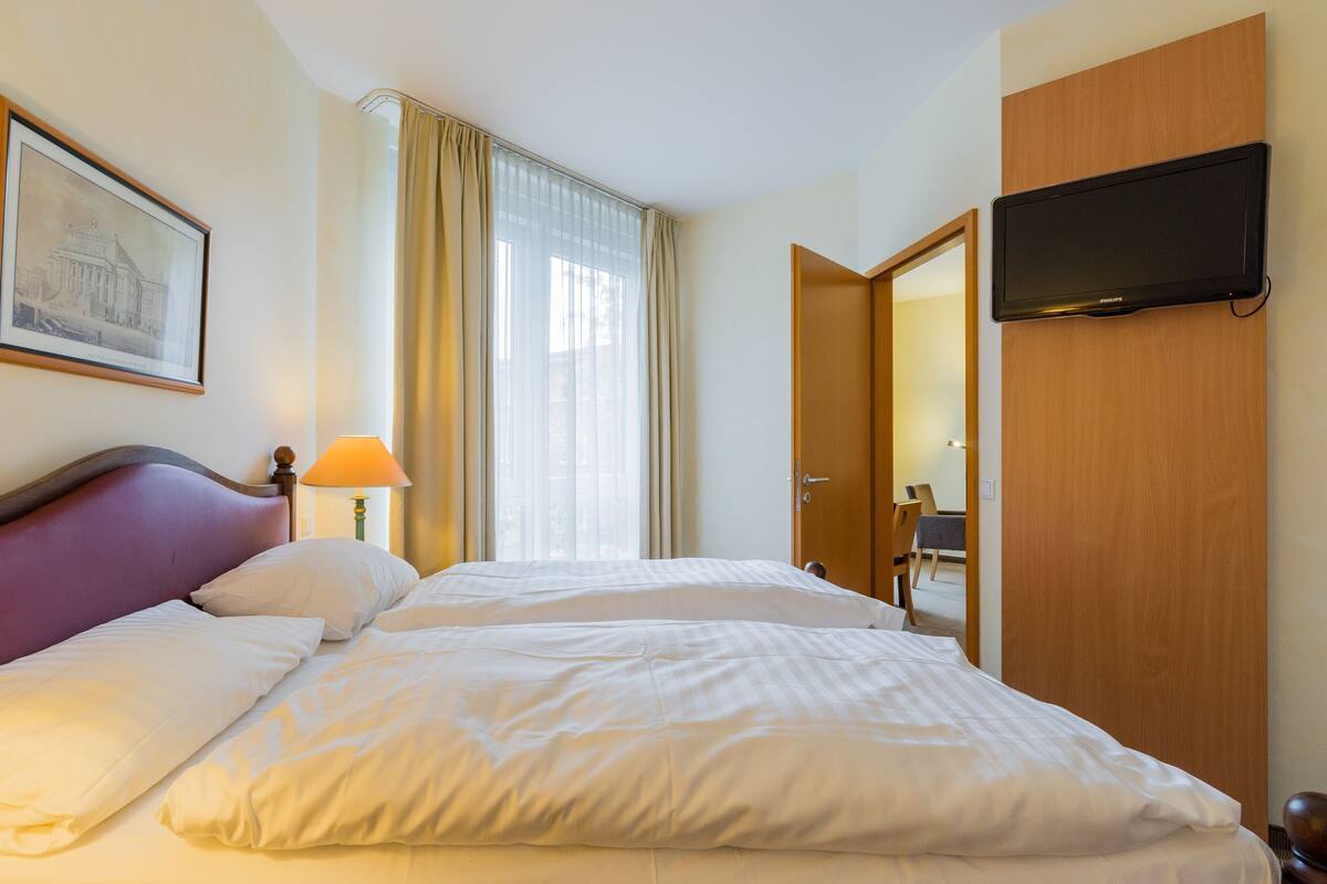 A hotel room with a bed, a television mounted on the wall, a framed picture on the wall, and a window with curtains.