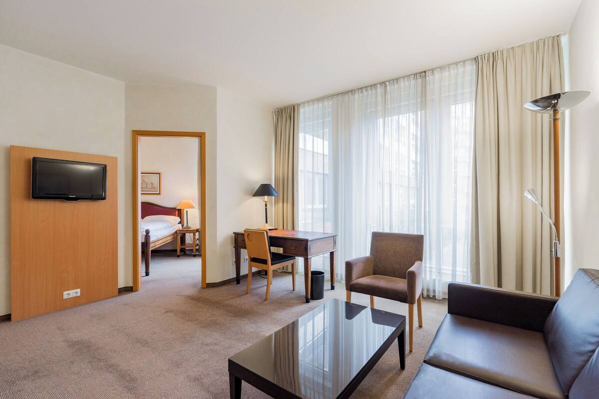 A hotel room with a bed, a desk, a chair, a sofa, a TV, and large windows with curtains.