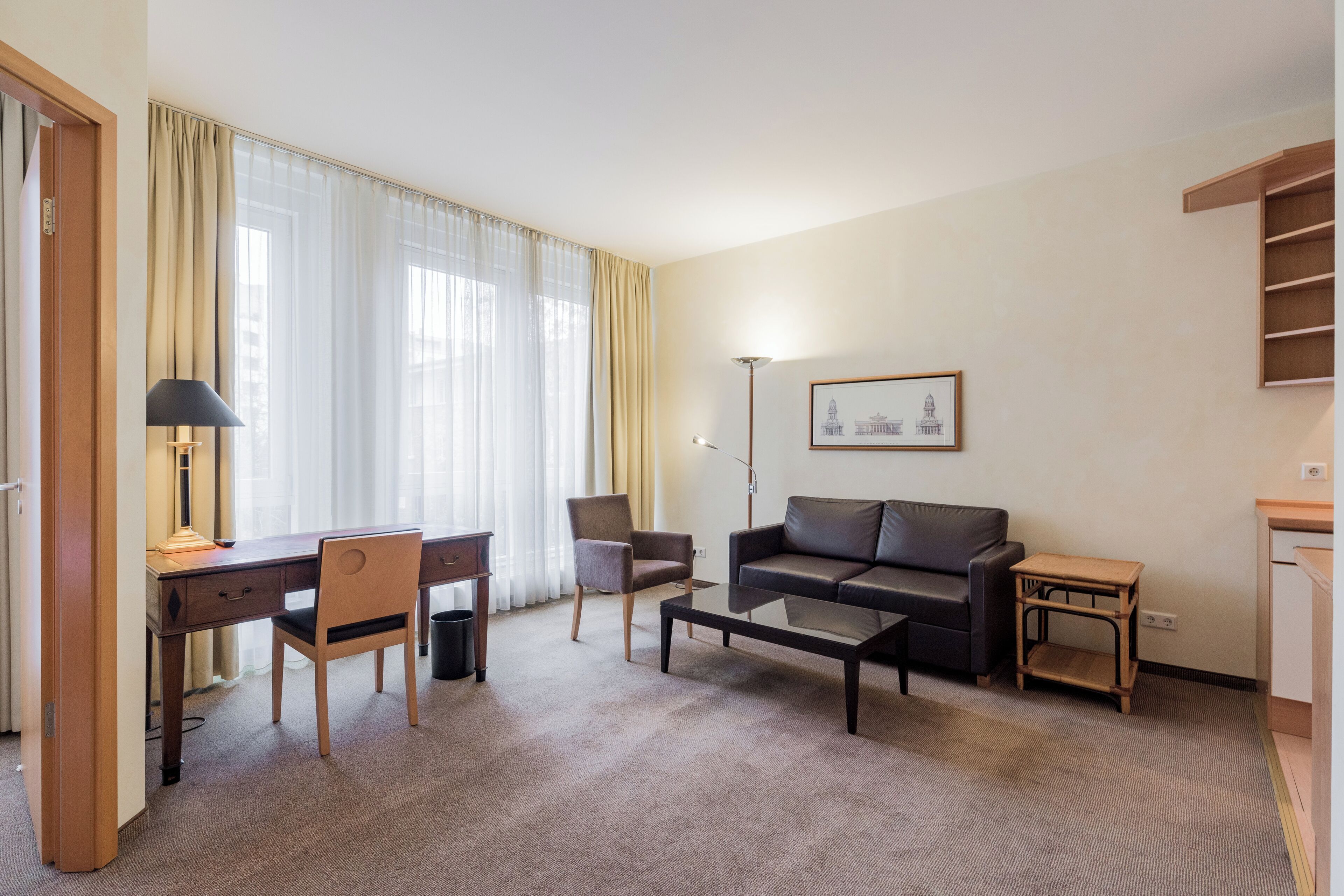 Quarto de hotel com uma escrivaninha, duas cadeiras, um sofá, uma mesa de centro, uma luminária, um quadro na parede e uma pequena mesa lateral de madeira.