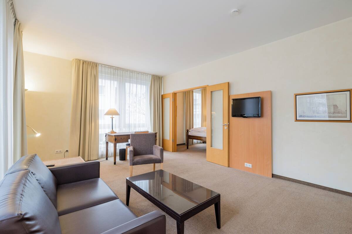 A hotel room with a sofa, a coffee table, a chair, a desk, a bed, a television, and a framed picture on the wall.