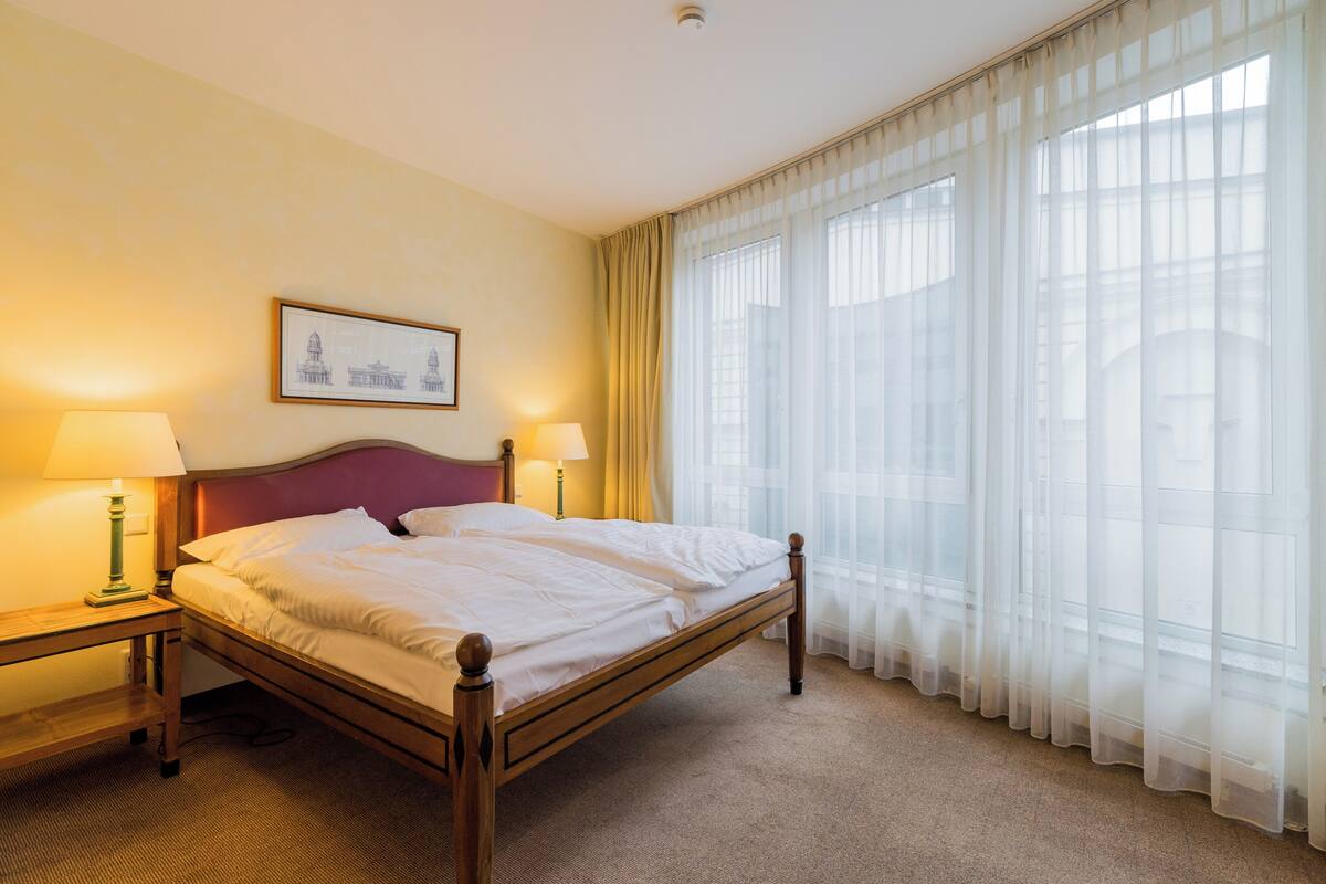 A hotel room with a wooden bed, two bedside lamps, a framed picture on the wall, and sheer curtains on the window.