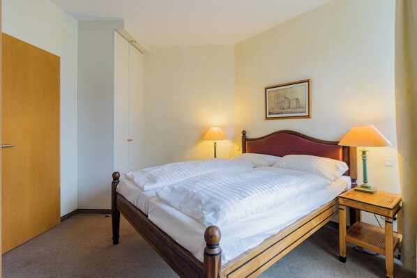 Un lit bien fait, avec du linge de lit blanc, dans une chambre d’hôtel. Une porte en bois se trouve à gauche et une table de chevet avec une lampe est placée à droite.