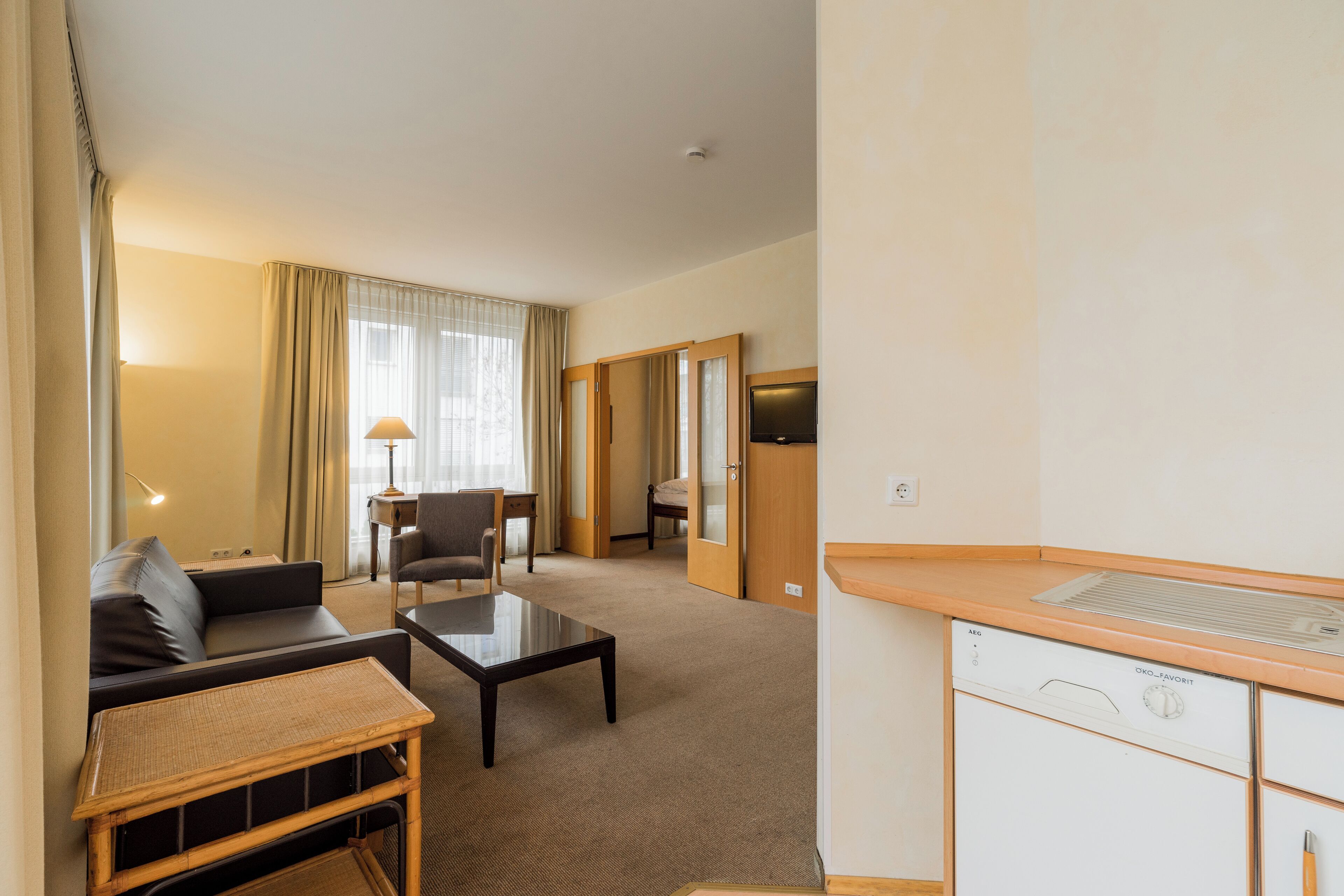 A hotel room with a sofa, a small table, a lamp, a television, and a kitchenette area.