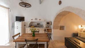 A compact kitchen with a microwave, oven, and a small refrigerator. There is a dining table with chairs and a vase of flowers. An arched stone wall is visible in the background.