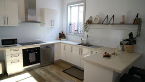 A modern kitchen with white cabinets, stainless steel appliances, a window with plants, and a small island with food and a glass. - Stay on Summer | CBD Apartment (Orange)