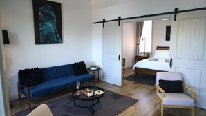 A modern room with a blue sofa, a small round table with a glass of wine, a wooden chair, a bed with white bedding, and a painting on the wall. - Stay on Summer | CBD Apartment (Orange)