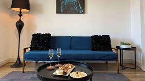 A living room with a blue sofa, a black floor lamp, a framed artwork of a parrot, and a coffee table with a plate of food and two glasses. - Stay on Summer | CBD Apartment (Orange)