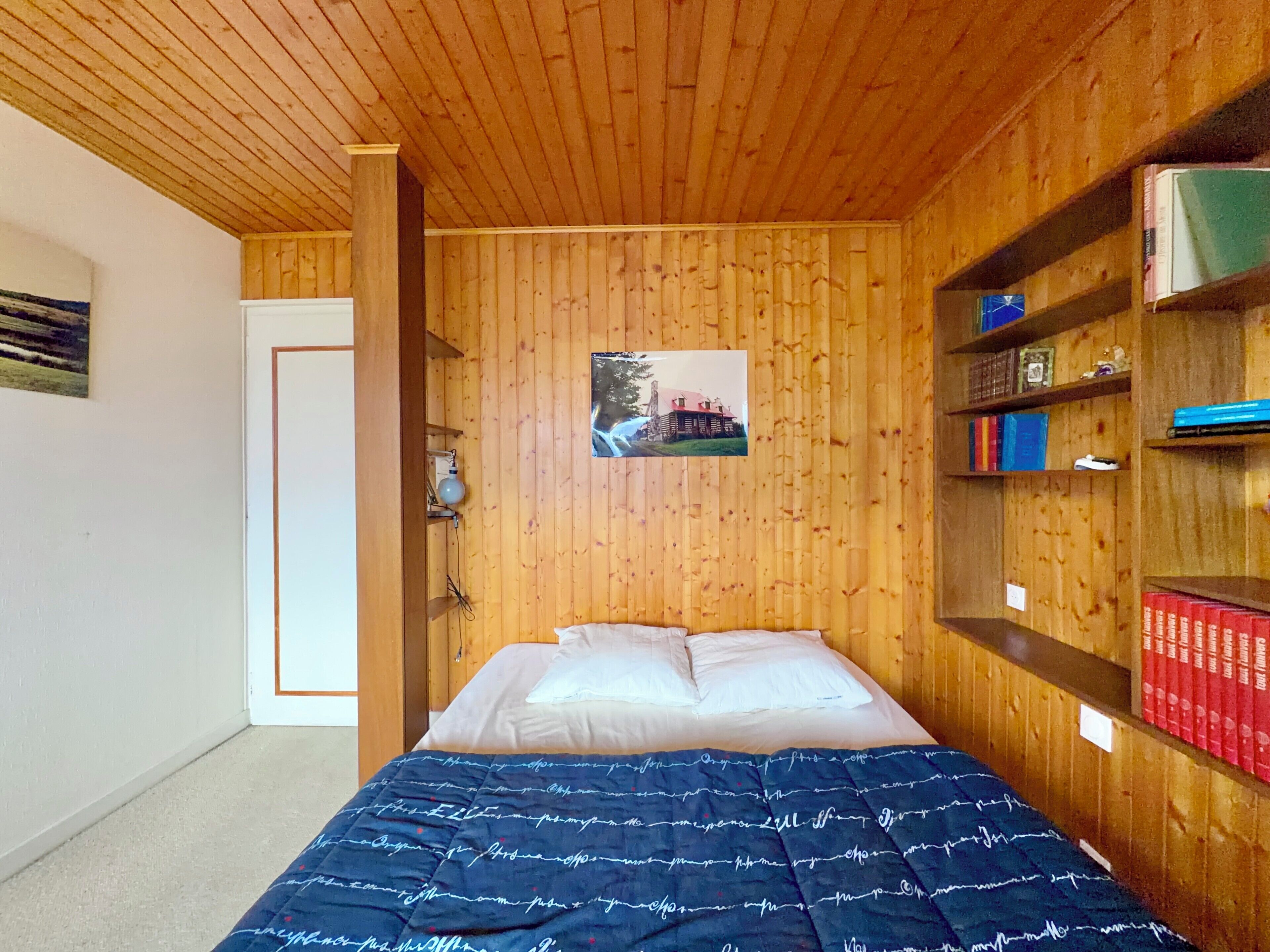 A bedroom with a wooden ceiling, a bed with white bedding, a blue patterned blanket, a wall-mounted shelf with books, and a framed picture on the wall.