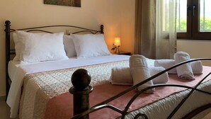 A hotel room with a bed, white linens, a painting on the wall, a nightstand with a lamp, and a window with curtains. - Stunning 3-bedroom Villa Near Malia Beach (Agios Nikolaos)