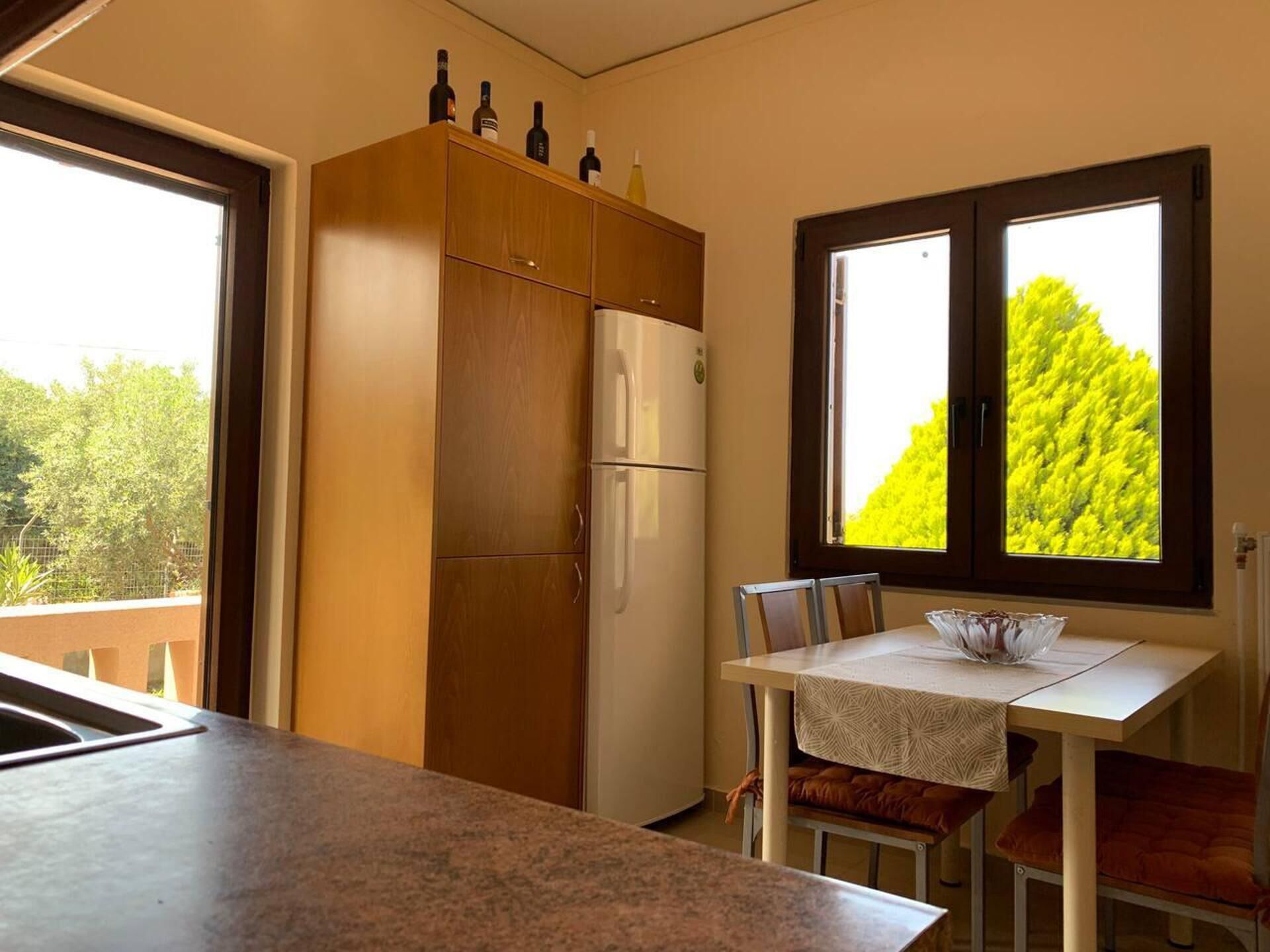 A kitchen with a refrigerator, a dining table with chairs, and a window with a view of trees.