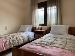 A room with two single beds, a wooden nightstand, a window with curtains, and a radiator. - Stunning 3-bedroom Villa Near Malia Beach (Agios Nikolaos)
