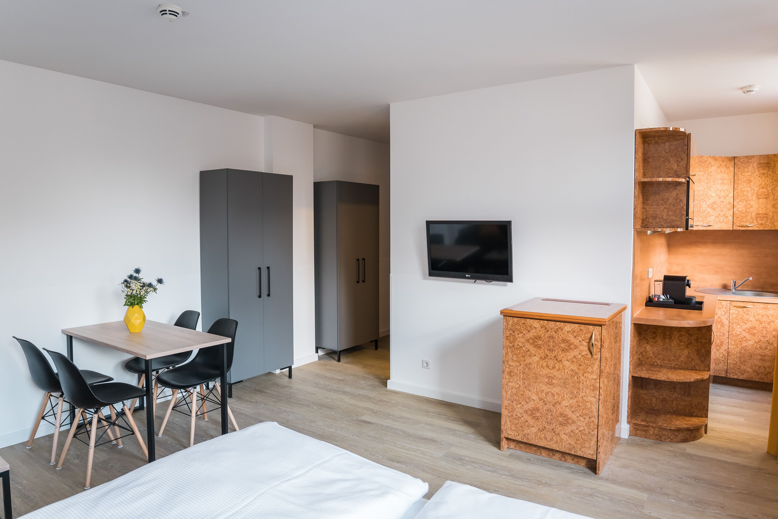 A compact hotel room with a bed, a dining table with chairs, a TV, and a kitchenette.