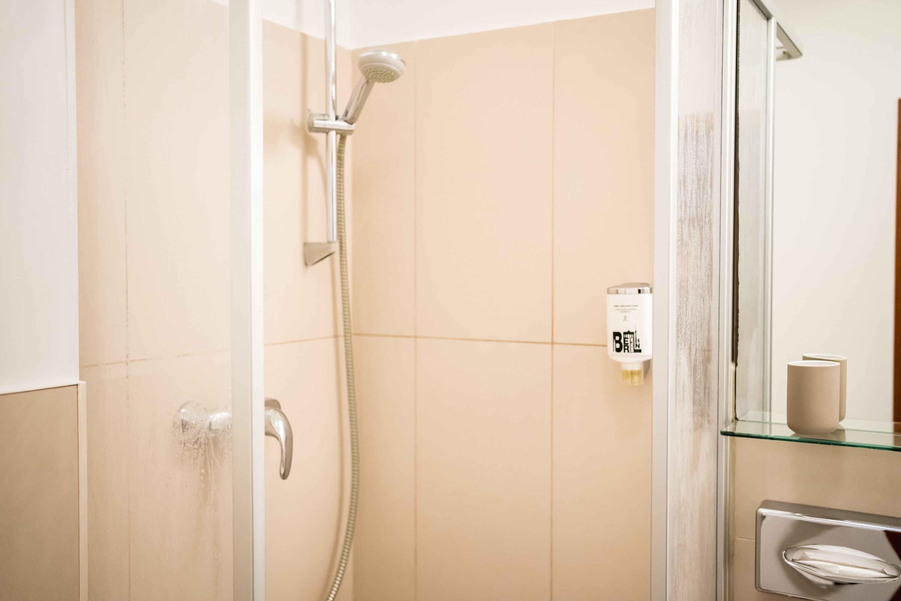 A modern bathroom with a glass shower enclosure, a handheld showerhead, and a visible soap dispenser on the wall.