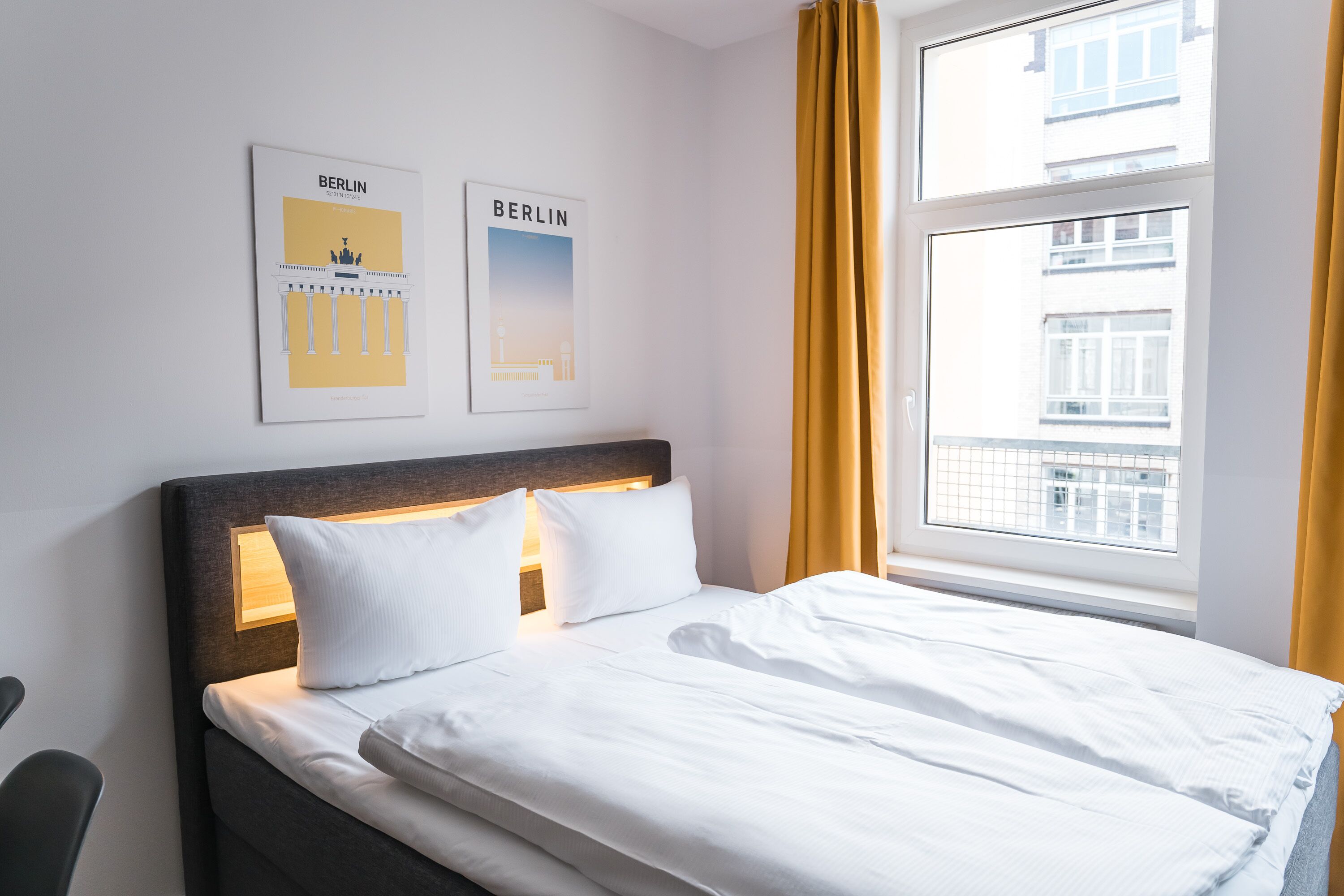 A hotel room with a bed, two framed pictures of Berlin, a window with curtains, and a view of buildings outside.
