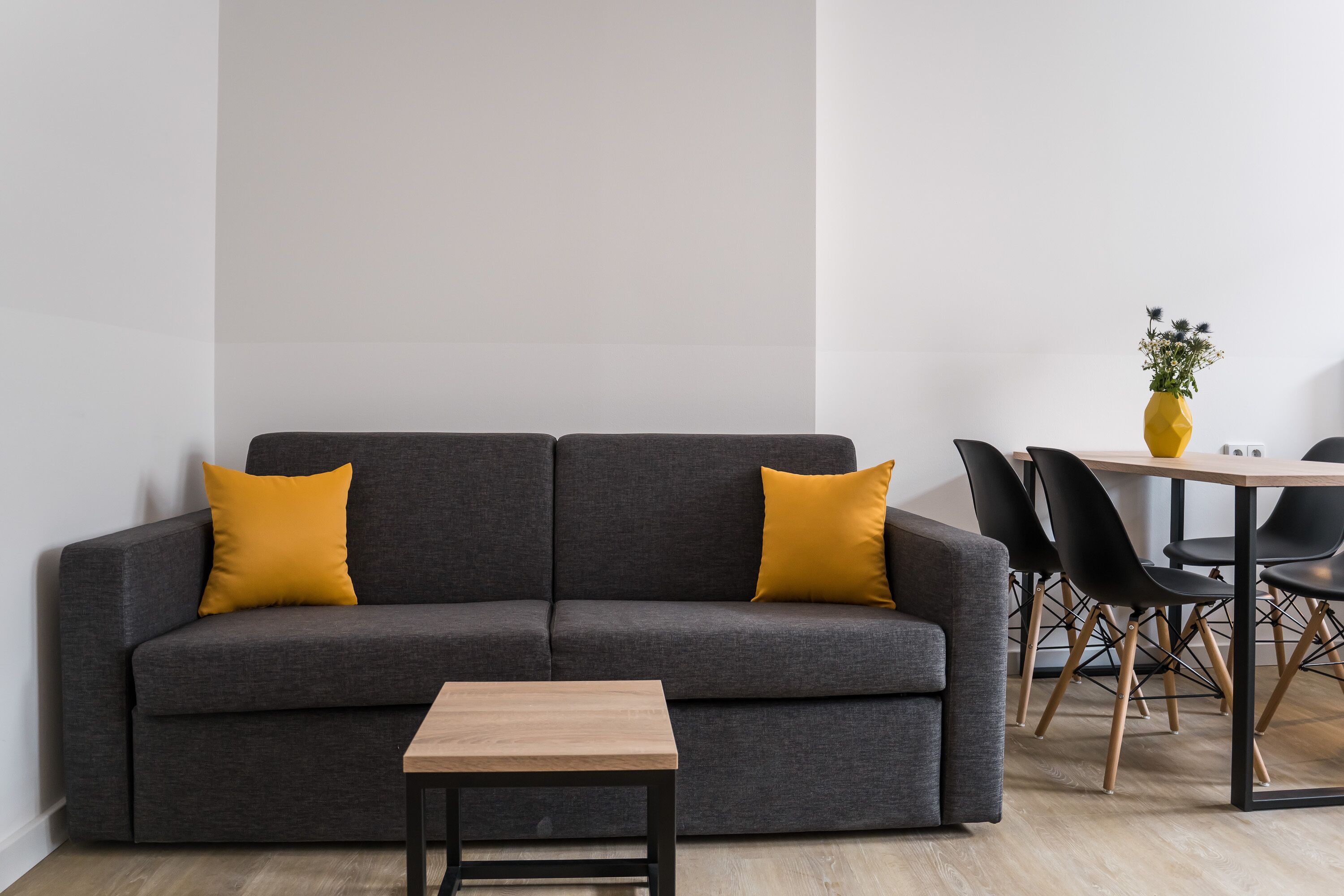 A modern living room with a grey sofa, two mustard-colored pillows, a small wooden table, and a dining area with a white table and black chairs.