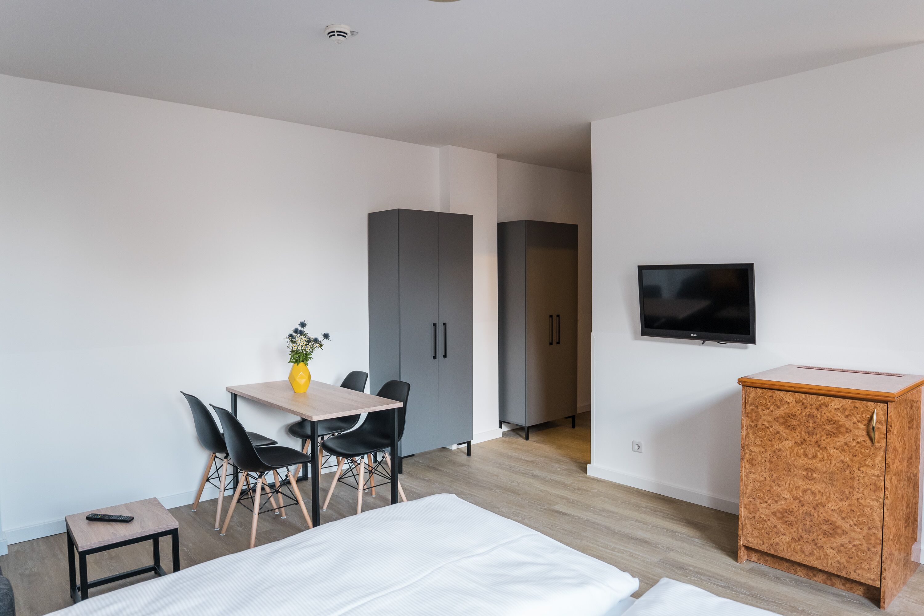 A modern hotel room with a bed, a desk with chairs, a small table with a vase, a TV on the wall, and a wardrobe.