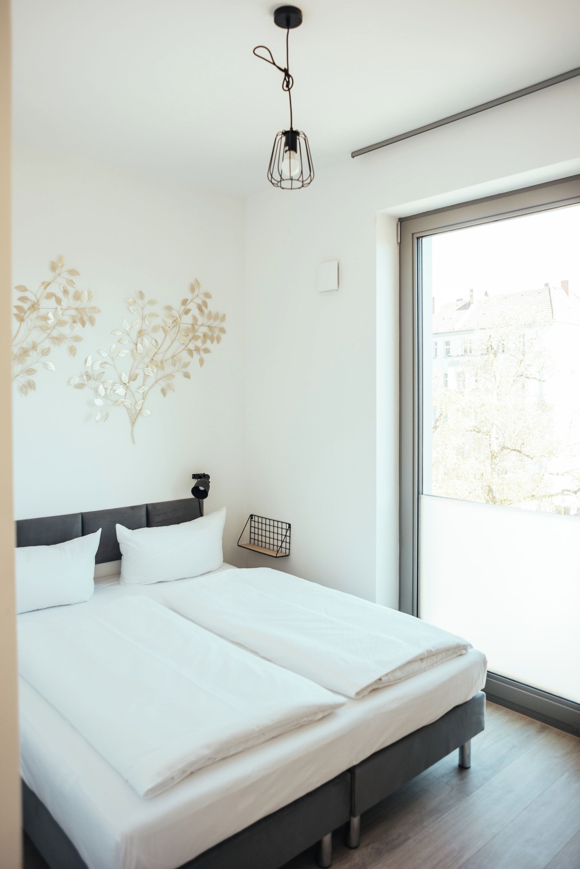 Un dormitorio moderno con una cama grande, una mesita de noche pequeña, una lámpara de pared y una ventana con vistas a edificios.